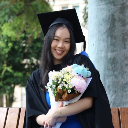 A young woman in graduation cap and gown sits on a bench seat in UQ's Great Court, clutching a bouquet of flowers with a small teddy bear inside it.
